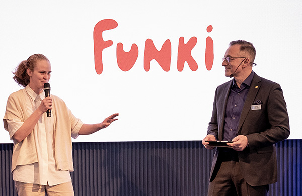 En kvinna pratar i en mikrofon på en scen. En man tittar på.